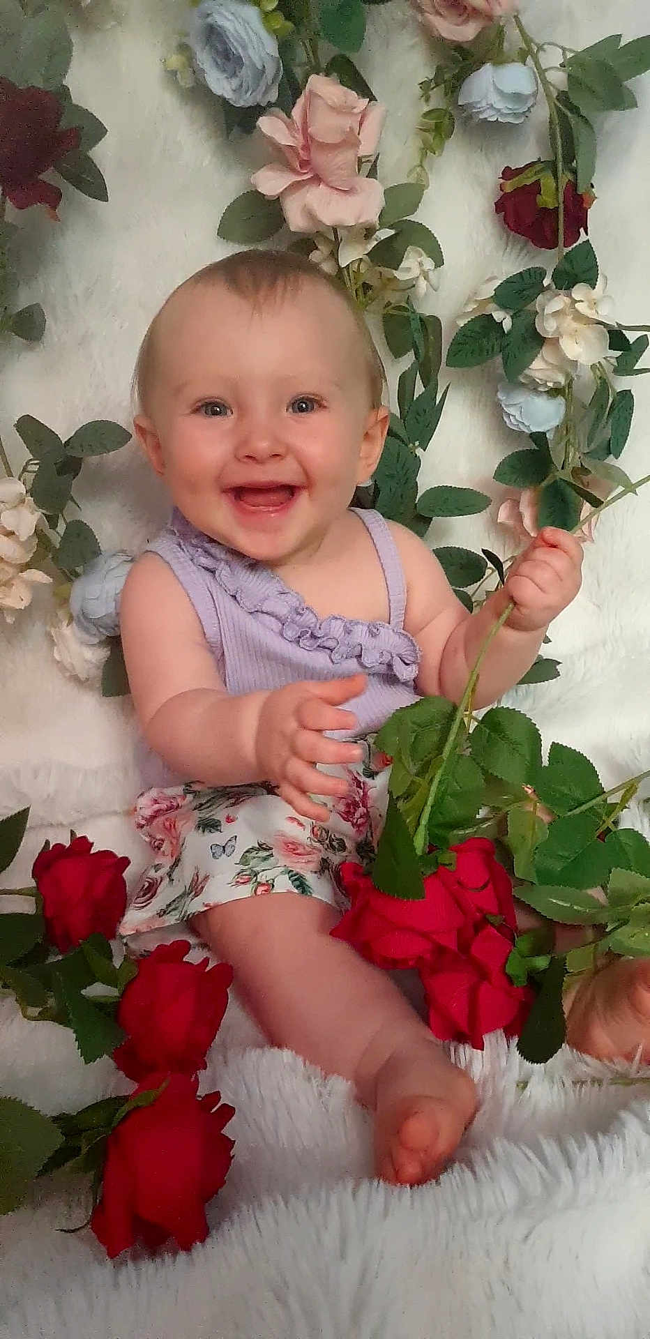 Ilona a rejoint le concours — aidez-le/la à gagner de superbes lots ! baby, smiling, flower, rose, purple_top, floral_skirt, green_leaves, sitting, happy, cute, infant, plant, petals, nature, portrait, child, cheerful, skin, barefoot, indoor