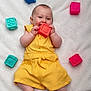 Ilona a rejoint le concours — aidez-le/la à gagner de superbes lots ! baby, child, yellow_outfit, toy_block, red_block, blue_block, pink_block, green_block, soft_blanket, smiling, chewing, happy, infant, playtime, colorful, cute, lying_down, indoors, skin, small_toys