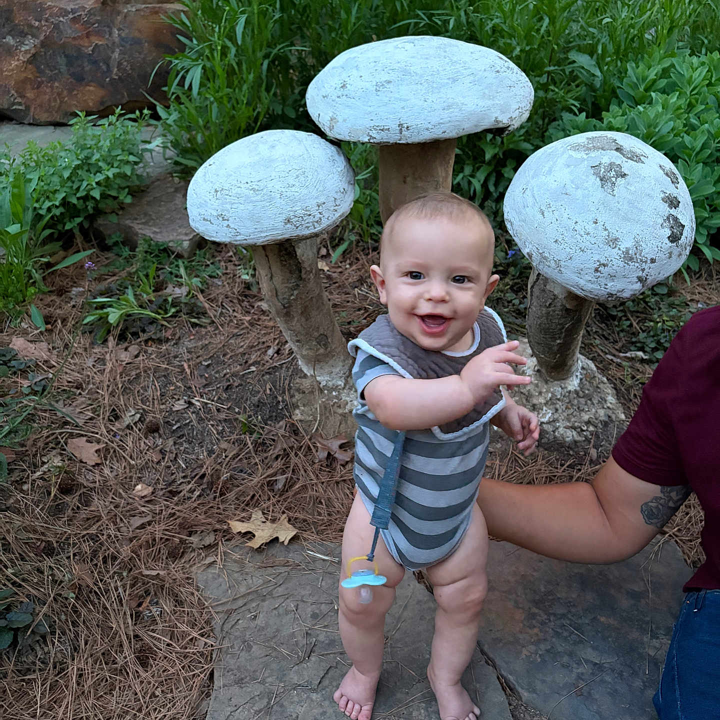 Colby joined the competition — help win amazing prizes! baby, child, smiling, outdoor, nature, mushrooms, greenery, stone, barefoot, onesie, bib, pacifier, tattoo, arm, person, plant, foliage, happy, standing, support