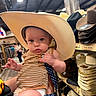 Colby is registered to the contest to win money with this photo: baby, child, cowboy_hat, striped_clothing, pacifier, store, hats, shelf, indoor, person_holding, curious_expression, aisle, wooden_floor, headwear, shopping, retail, infant, cute, hand, adult
