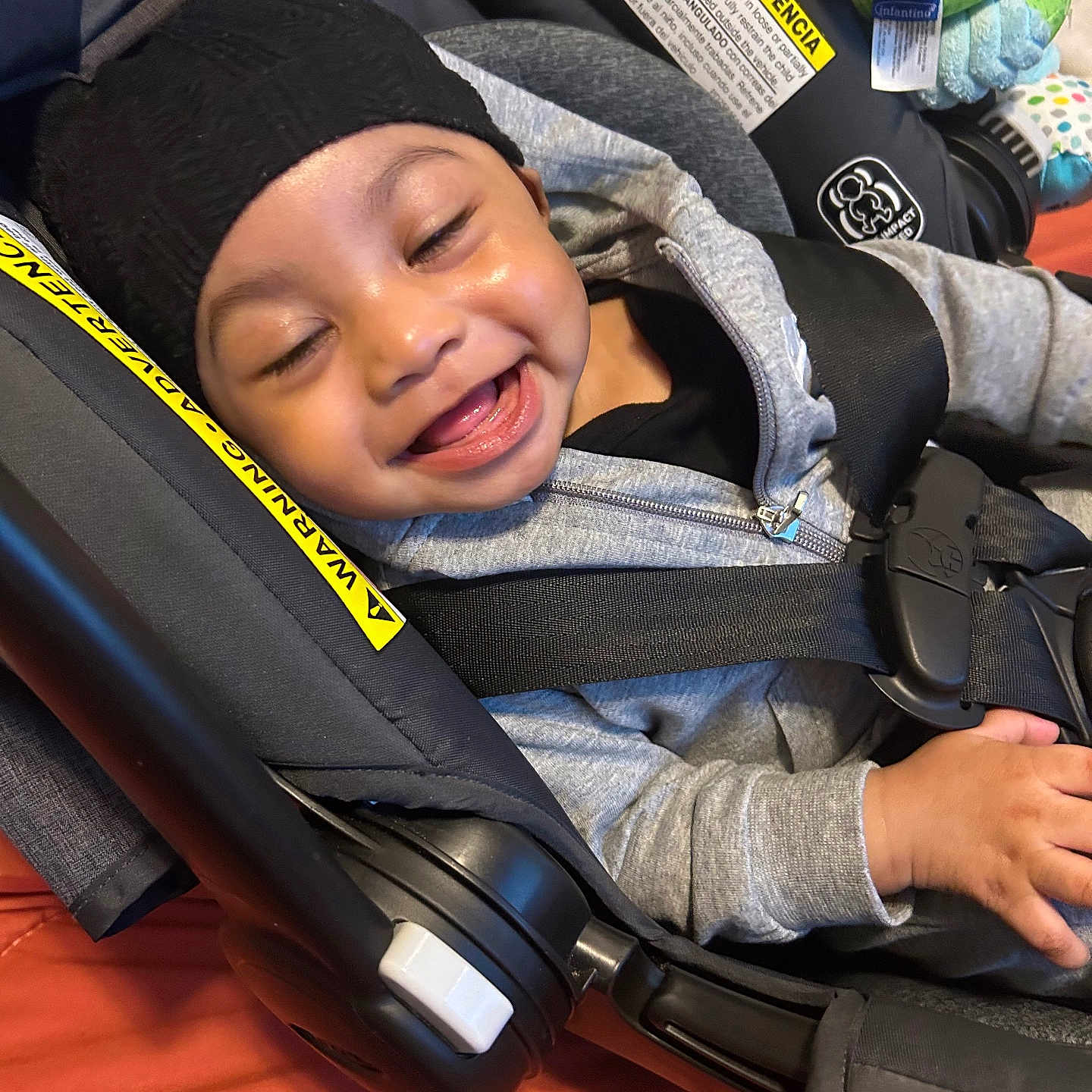 Ryan is registered to the contest to win money with this photo: baby, child, infant, smiling, laughing, portrait, stroller, car_seat, safety_harness, black_hat, gray_hoodie, plush_toy, toy, seated, indoor, close_up, happy, face, transportation, photography