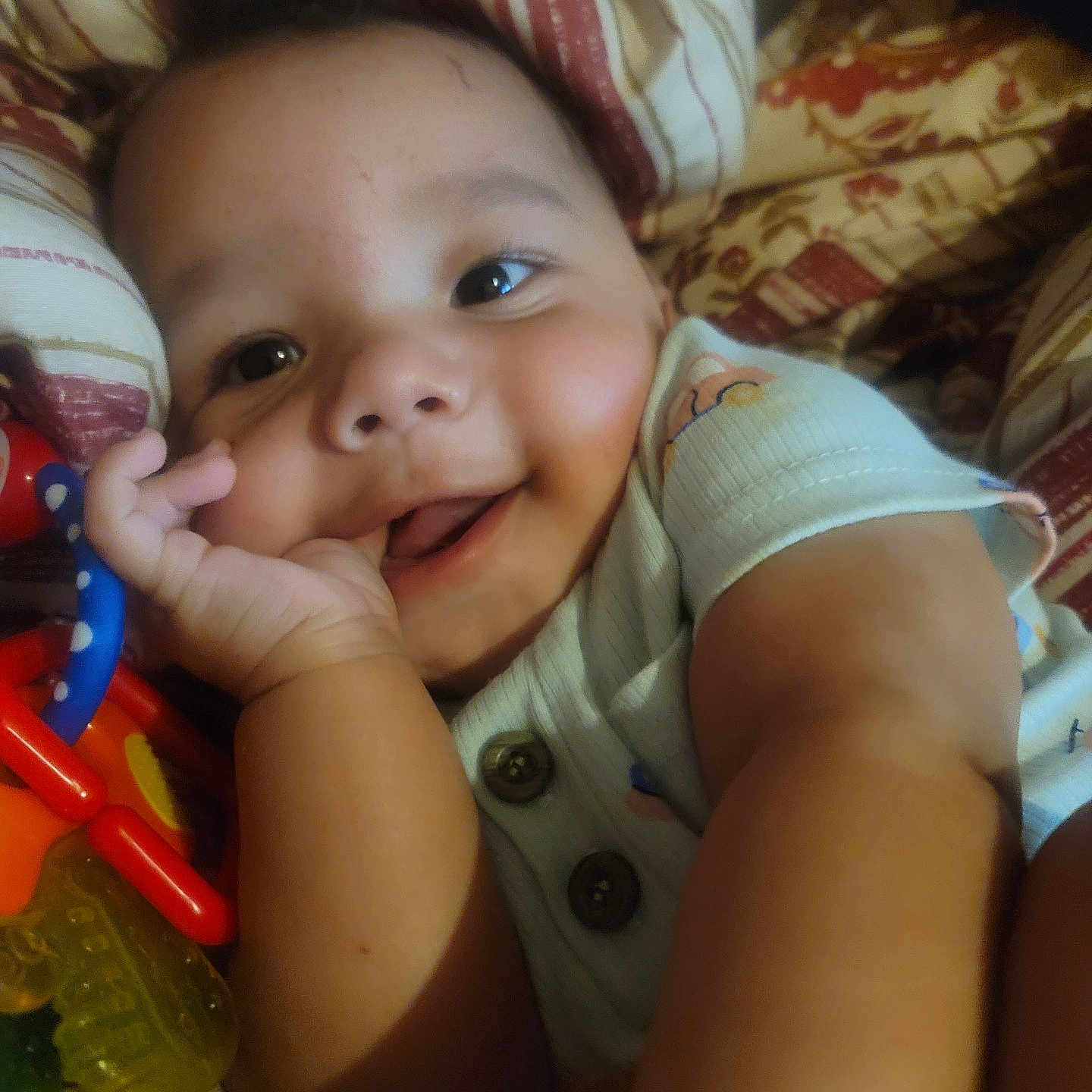 Daxton is registered to the contest to win money with this photo: baby, smiling, hand, face, toy, blanket, pattern, child, infant, happy, cute, skin, clothing, closeup, person, lying_down, indoors, colorful, comfort, portrait