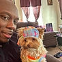 man, dog, pet, indoor, living_room, couch, curtains, tie_dye, hat, cute, small_dog, portrait, smiling, casual, furniture, carpet, human, colorful, window, relaxing