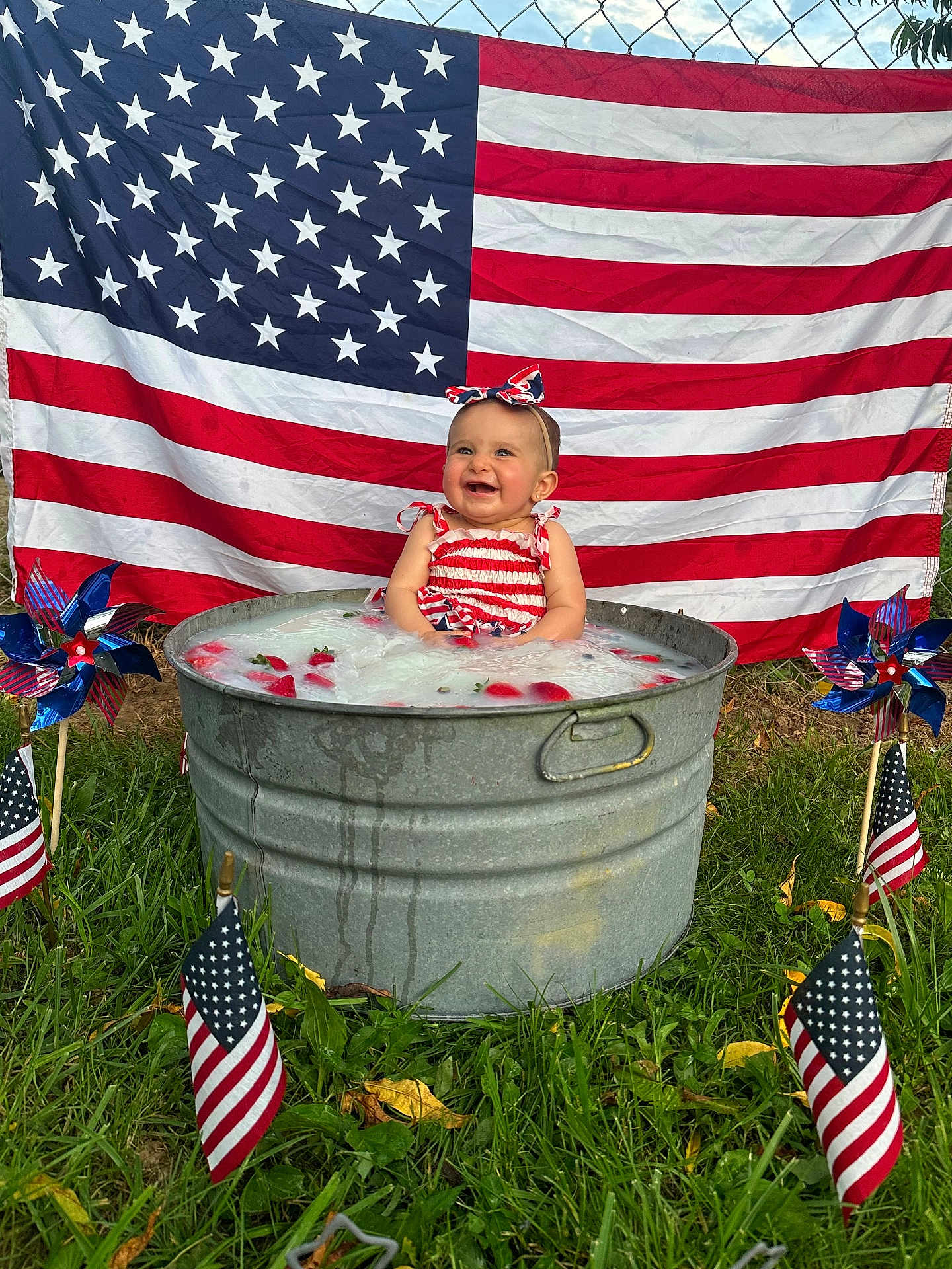 Stella is registered to the contest to win money with this photo: baby, child, american_flag, metal_tub, strawberries, milky_water, grass, pinwheel, small_flags, headband, outdoor, summer, festive, red_white_blue, happy, smiling, portrait, patriotic, nature, celebration