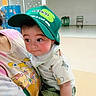 baby, child, person, cap, hat, green_cap, tshirt, cartoon_print, indoor, locker, cart, floor, curious, holding, shorts, clothing, headwear, cute, face, human