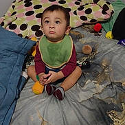 Tobias is registered to the contest to win money with this photo: toddler, child, baby, bed, blanket, pillow, toy, bib, socks, indoor, cute, cross_legged, expression, looking_up, patterned, colorful, soft_toys, casual_clothing, floor_bedroom, home