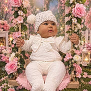 Tiffany is registered to the contest to win money with this photo: baby, child, face, fairy_tale, floral_decoration, flowers, garden, hands, infant, knit_hat, outdoor, photography, pink_flowers, pom_pom, portrait, roses, sitting, smiling, swing, white_outfit