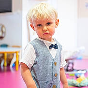 Rémy a rejoint le concours — aidez-le/la à gagner de superbes lots ! background, blond_hair, bow_tie, casual, child, clothing, colorful, floor, furniture, indoor, person, playroom, portrait, serious_expression, standing, toddler, toy, vest, white_shirt, young_child
