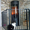 cat, black_and_white_cat, fireplace, fire, safety_gate, tile_floor, stone_wall, wood_box, indoor, pet, animal, yellow_eyes, furniture, home_interior, cozy, warm, sitting, domestic_cat, glass, modern