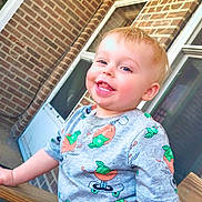 Myles is registered to the contest to win money with this photo: toddler, child, baby, smiling, happy, portrait, outdoor, brick_wall, house, window, door, wooden_railing, t_shirt, patterned_clothing, orange_pattern, skateboard_pattern, red_shorts, standing, daylight, close_up