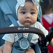 Maryam joined the competition — help win amazing prizes! baby, child, stroller, headband, bow, face, eyes, pout, clothing, seatbelt, infant, person, outdoor, blurred_background, baby_seat, baby_clothes, hand, straps, closeup, portrait