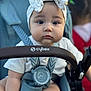 baby, child, stroller, headband, bow, face, eyes, pout, clothing, seatbelt, infant, person, outdoor, blurred_background, baby_seat, baby_clothes, hand, straps, closeup, portrait