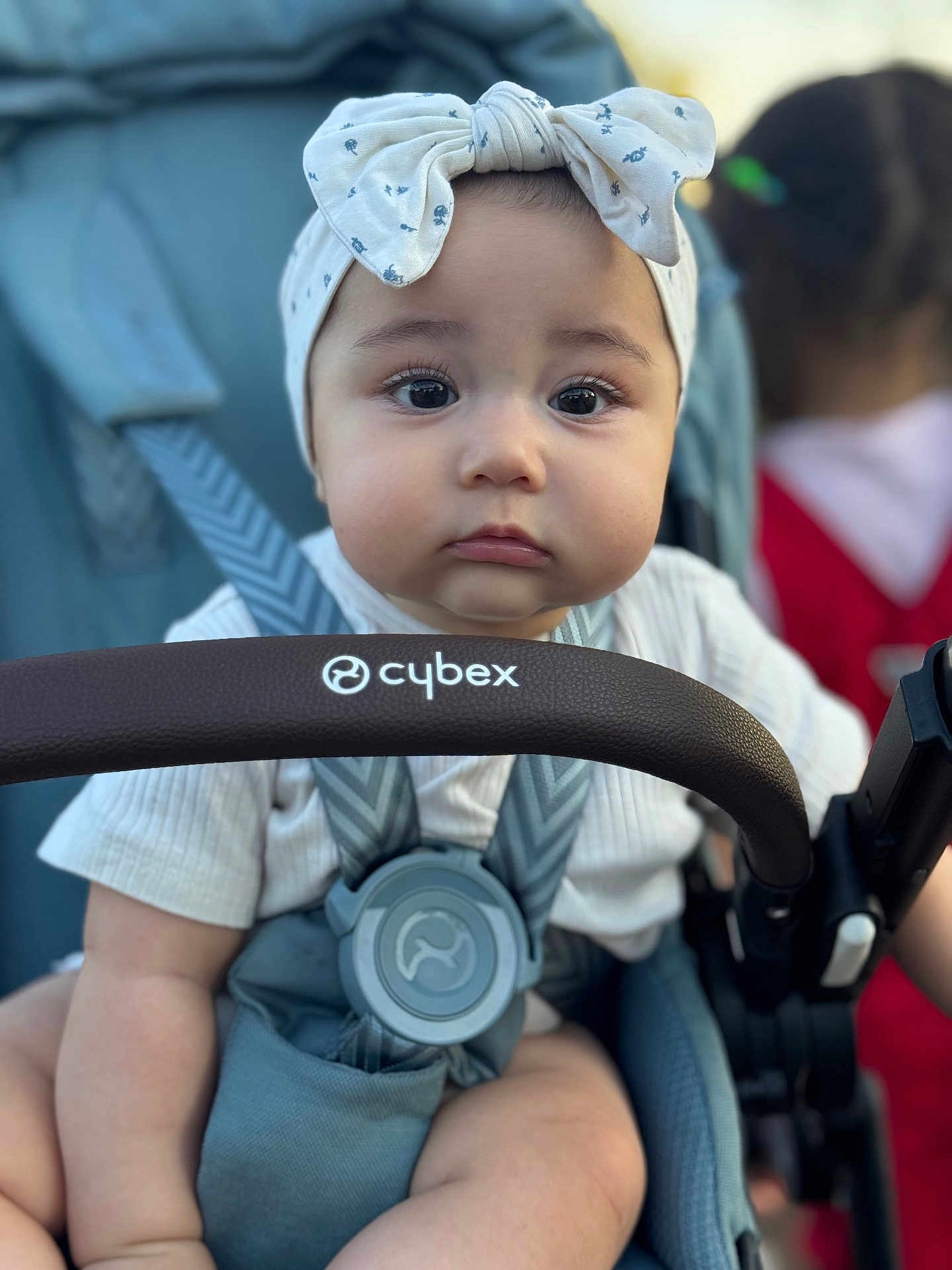 Maryam joined the competition — help win amazing prizes! baby, child, stroller, headband, bow, face, eyes, pout, clothing, seatbelt, infant, person, outdoor, blurred_background, baby_seat, baby_clothes, hand, straps, closeup, portrait