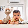 baby, child, plush_toy, stuffed_animal, bed, pillow, blanket, indoor, curious, soft_toy, window, frame, photograph, home, cute, infant, person, casual_clothing, face, lying_down