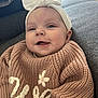 apparel, baby, bow, child, couch, cozy, cute, face, happy, headband, indoor, infant, knitted_sweater, person, portrait, relaxed, sitting, smiling, warm_clothing, young_child