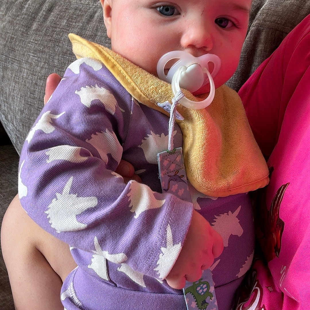 Riley is registered to the contest to win money with this photo: baby, bib, blue_eyes, child, comfort, couch, cute, fabric, face, hair, hand, indoor, infant, onesie, pacifier, person_holding_baby, portrait, purple_clothing, soft_light, unicorn_pattern