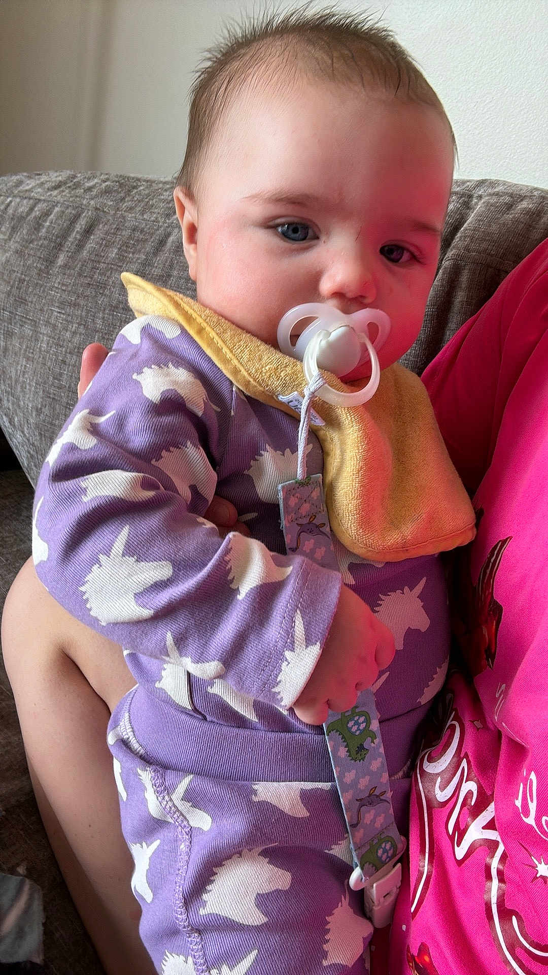 Riley is registered to the contest to win money with this photo: baby, infant, pacifier, onesie, purple_clothing, unicorn_pattern, bib, couch, person_holding_baby, hand, face, blue_eyes, hair, indoor, soft_light, fabric, comfort, child, cute, portrait