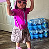 child, toddler, girl, portrait, standing, umbrella, pink, sunglasses, hat, fashion, skirt, sandals, indoor, home, wood_floor, door, water_bottles, smiling, pose, colorful