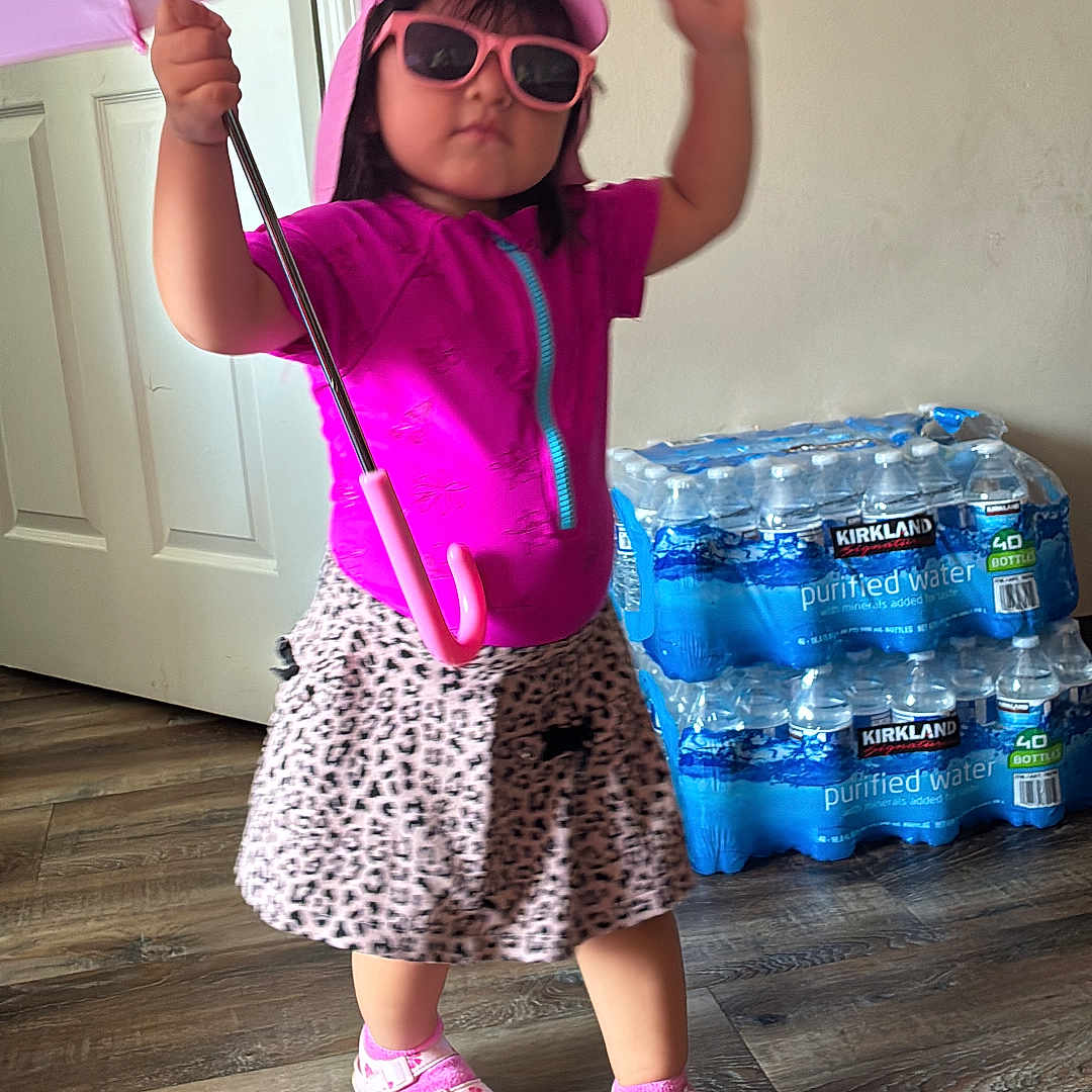 Raisha joined the competition — help win amazing prizes! child, toddler, girl, portrait, standing, umbrella, pink, sunglasses, hat, fashion, skirt, sandals, indoor, home, wood_floor, door, water_bottles, smiling, pose, colorful