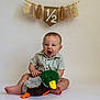 Clayton joined the competition — help win amazing prizes! baby, child, plush_toy, duck, sitting, milestone, banner, tassels, happy, cute, indoor, portrait, smiling, striped_shirt, feet, floor, celebration, anniversary, one_half, expression