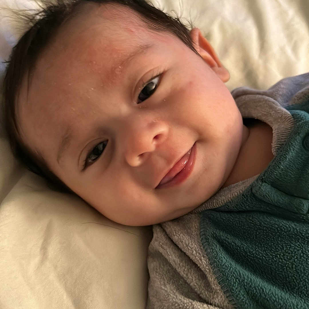 Noah Smith Pérez is registered to the contest to win money with this photo: baby, blanket, child, closeup, clothing, cozy, cute, eyelashes, face, hair, happy, head, indoors, infant, lying_down, portrait, skin, smiling, soft_texture, young_child