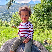 Fael a rejoint le concours — aidez-le/la à gagner de superbes lots ! child, toddler, curly_hair, striped_shirt, rock, outdoor, nature, mountains, greenery, grass, tree, sunny, smiling, casual_clothing, sneakers, happy, portrait, daylight, scenic, playful