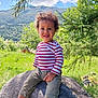 child, toddler, curly_hair, striped_shirt, rock, outdoor, nature, mountains, greenery, grass, tree, sunny, smiling, casual_clothing, sneakers, happy, portrait, daylight, scenic, playful
