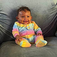 Marisol is registered to the contest to win money with this photo: baby, child, smiling, onesie, rainbow_colors, sitting, couch, gray_couch, hand, adult_hand, happy, indoor, person, furniture, soft_texture, portrait, cute, young_child, cozy, bright_colors