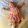 Marisol is registered to the contest to win money with this photo: baby, child, infant, dress, headband, bow, smile, lying_down, bed, blanket, footwear, white_shoes, socks, curly_hair, bracelet, colorful, patterned_clothing, happy, cute, indoors