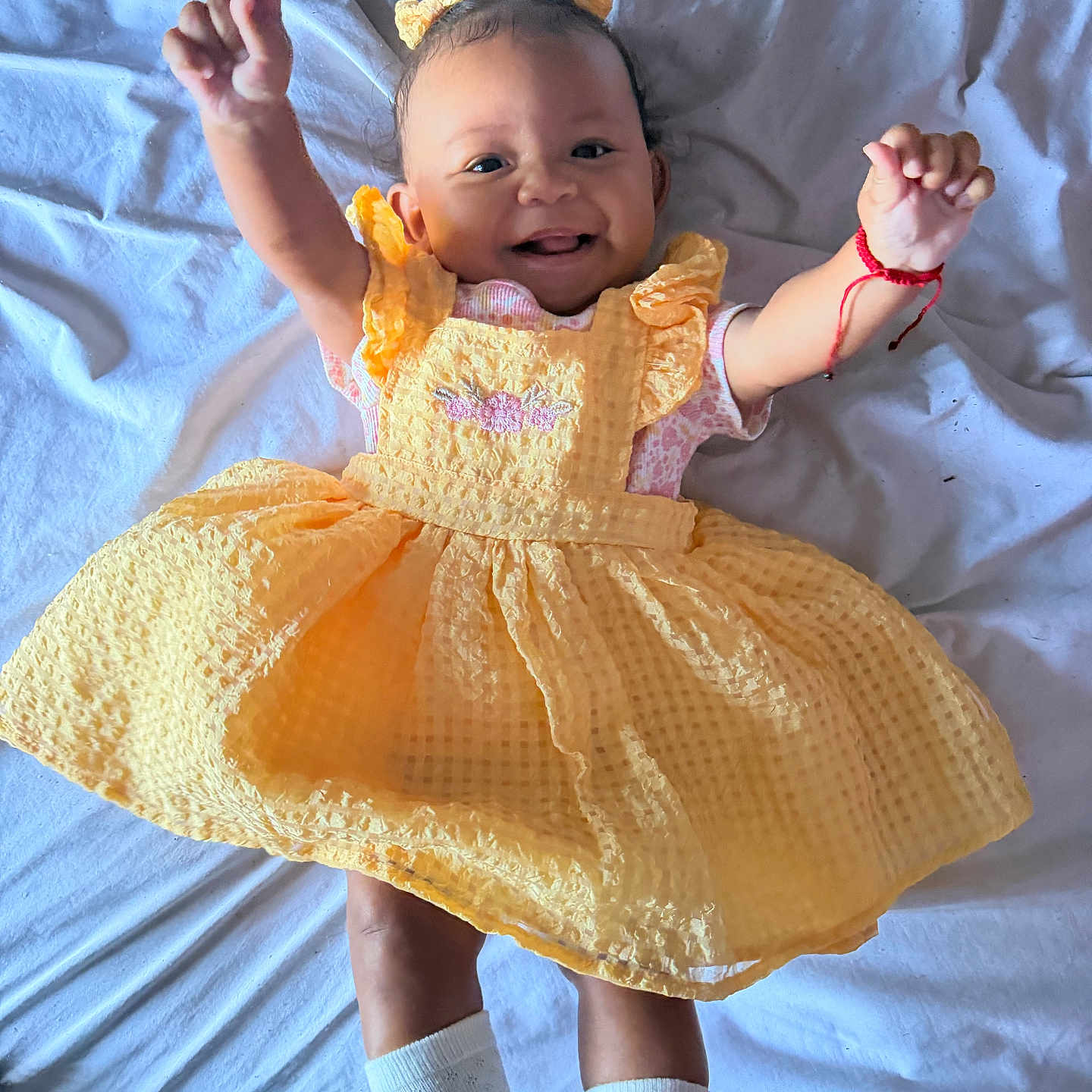 Marisol is registered to the contest to win money with this photo: baby, child, smile, yellow_dress, hair_bows, white_socks, lying_down, bed_sheet, happy, infant, portrait, cute, raised_arms, fabric_texture, indoors, person, skin, face, clothing, playful