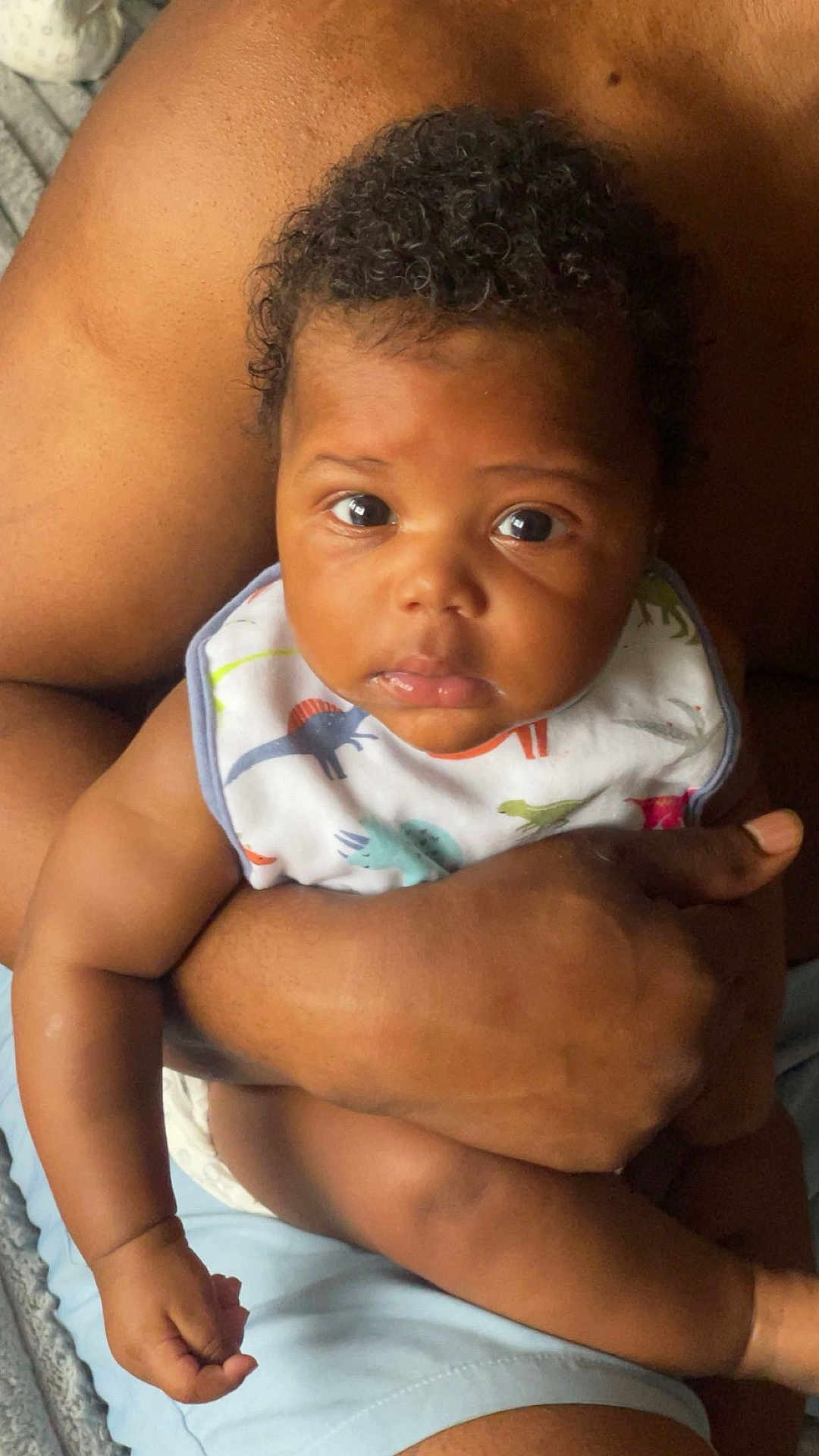 Kyaire is registered to the contest to win money with this photo: baby, infant, child, person, arm, hand, curly_hair, bib, dinosaur_pattern, skin, close_up, comfort, protection, expression, wide_eyes, indoor, holding, adult, caring, toddler