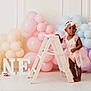 toddler, child, birthday, pink_dress, headband, balloons, pastel_colors, ladder, letters, one, celebration, indoor, portrait, smiling, decorations, party, festive, floor, white_background, cute