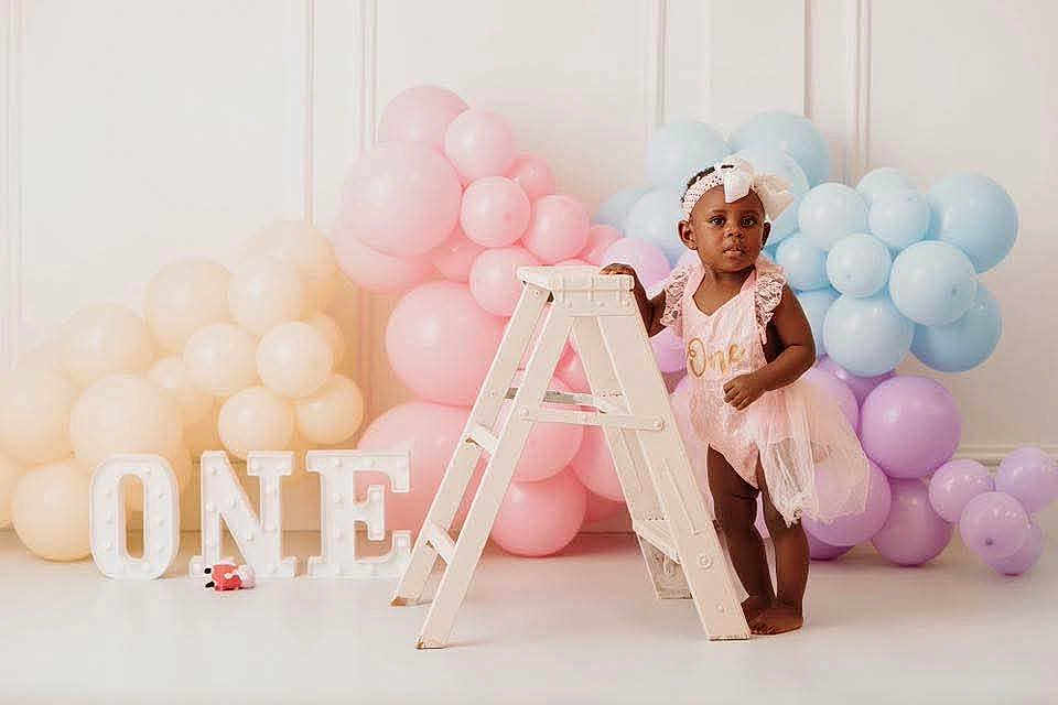 Leilani is registered to the contest to win money with this photo: toddler, child, birthday, pink_dress, headband, balloons, pastel_colors, ladder, letters, one, celebration, indoor, portrait, smiling, decorations, party, festive, floor, white_background, cute