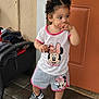 toddler, child, standing, minnie_mouse, clothing, shorts, tshirt, shoes, door, outdoor, pavement, curly_hair, hand, face, expression, casual, young, person, looking_away, footwear