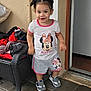 toddler, child, girl, smiling, standing, pigtails, minnie_mouse, tshirt, shorts, sneakers, wicker_chair, clothes, outdoor, doorway, concrete_floor, happy, cute, casual_clothing, person, daylight