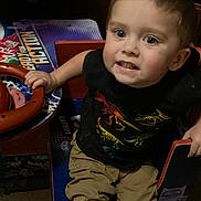 Jaxon is registered to the contest to win money with this photo: child, toddler, boy, face, eyes, hair, dinosaur_shirt, black_shirt, khaki_pants, hand, steering_wheel, arcade_machine, chair, indoor, carpet, portrait, candid, expression, smile, seat