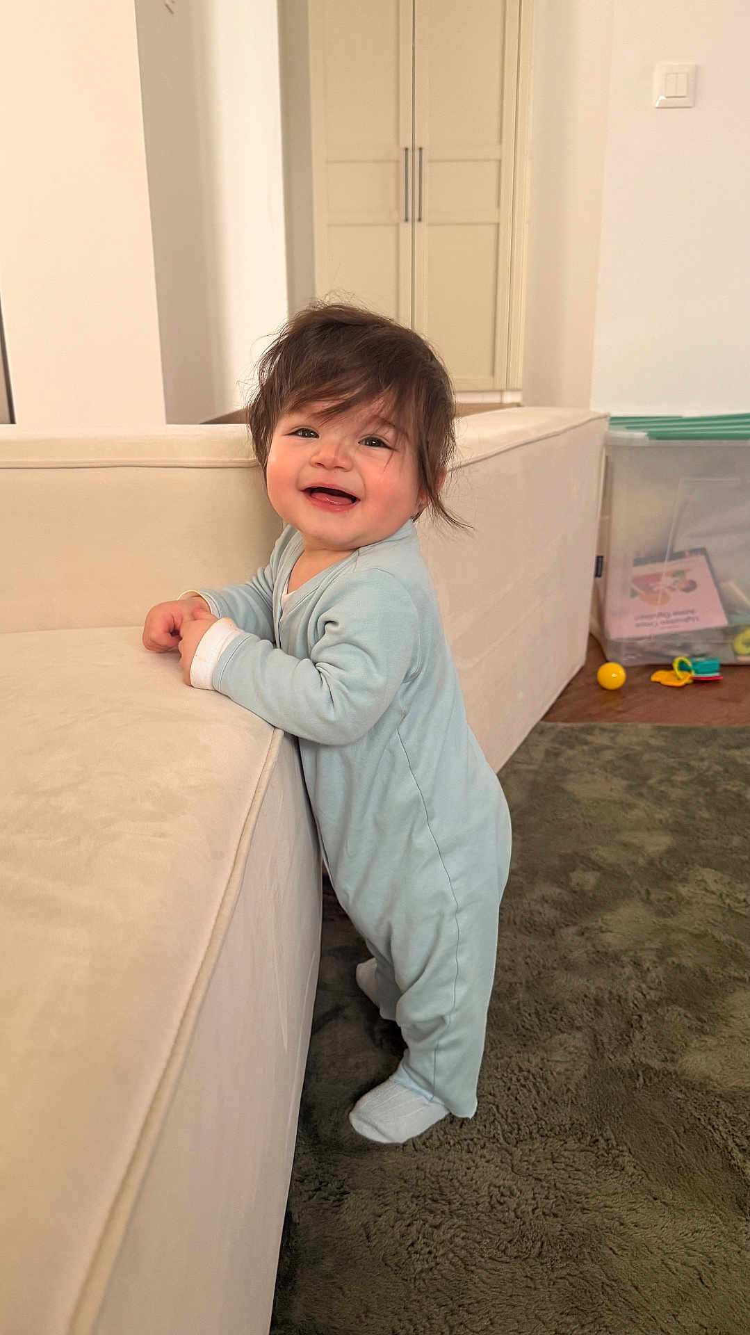 Aslan participe au concours pour gagner de l'argent avec cette photo : baby, child, onesie, standing, smiling, hair, couch, living_room, carpet, toy, plastic_box, floor, furniture, happy, indoor, person, cute, infant, home, play