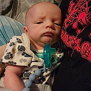 Sylus is registered to the contest to win money with this photo: baby, infant, pacifier, onesie, paw_prints, person, black_shirt, red_graphics, beaded_clip, quilted_background, closeup, indoor, holding, resting, cute, child, newborn, expression, soft_lighting, portrait