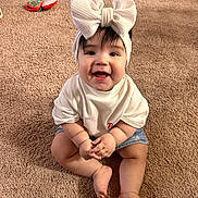 Bella is registered to the contest to win money with this photo: baby, child, smiling, headband, bow, white_shirt, denim_shorts, carpet, indoor, cute, happy, sitting, infant, person, skin, face, hands, toes, playful, young