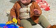 Haisley is registered to the contest to win money with this photo: toddler, child, sunglasses, toy, bed, pillow, plush_toy, brown_cardigan, green_pants, soft_shoes, indoor, playtime, cute, smiling, baby, patterned_bedspread, colorful_toy, happy, person, young_child