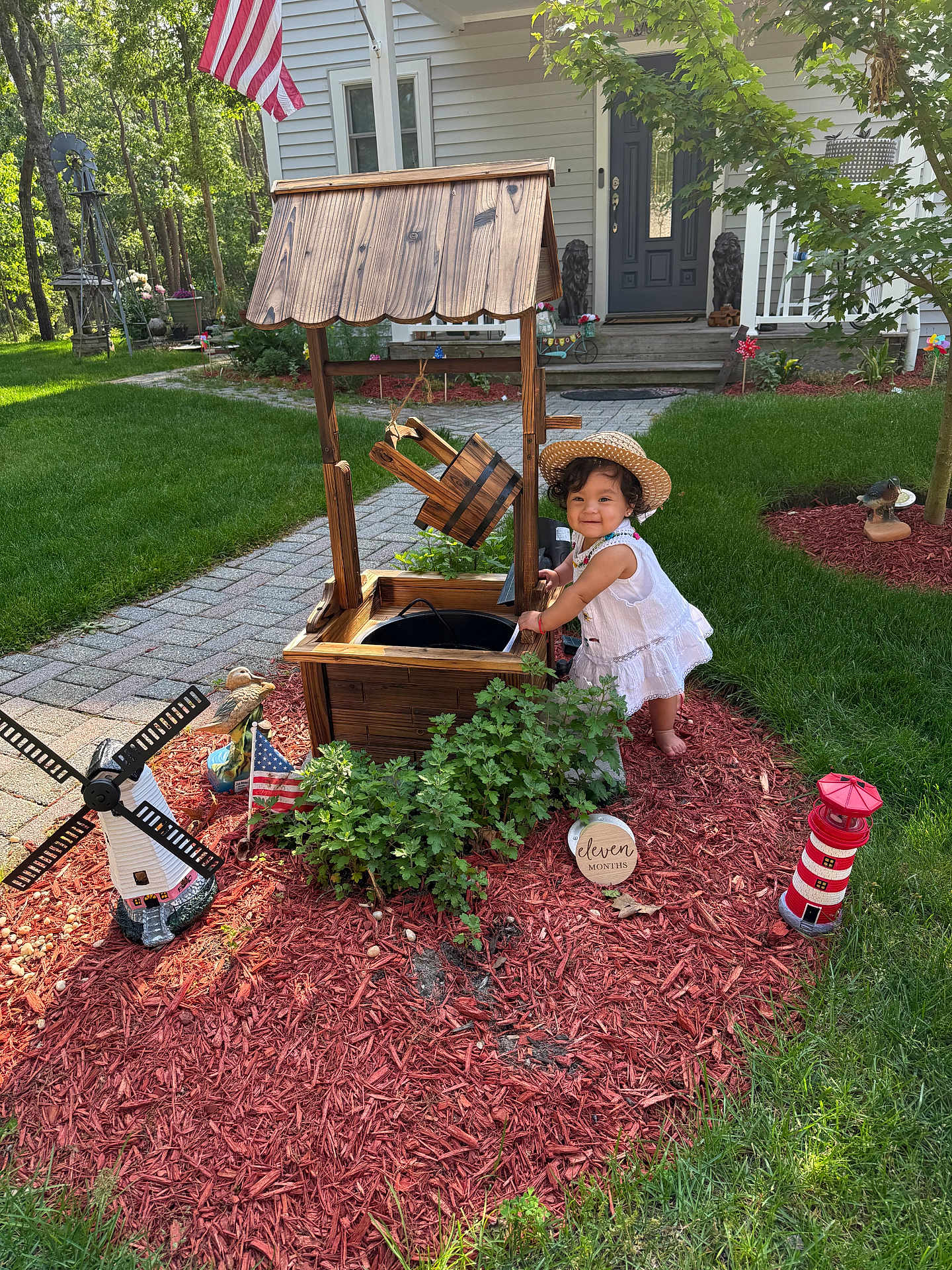 Haisley joined the competition — help win amazing prizes! baby, child, garden, mulch, wishing_well, wooden_structure, straw_hat, white_dress, grass, house, sidewalk, miniature_windmill, miniature_lighthouse, american_flag, plants, shrub, outdoor, sunlight, smiling, decorations