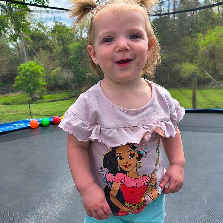 Trenity is registered to the contest to win money with this photo: toddler, child, pigtails, smiling, trampoline, outdoor, basketball_hoop, greenery, shorts, shirt, cartoon_character, daylight, play, happy, nature, grass, trees, person, young_child, summer_clothing