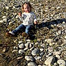 Avianna is registered to the contest to win money with this photo: child, toddler, rocks, riverbank, water, nature, outdoor, casual_clothing, jeans, sweater, boots, curly_hair, sunlight, shadow, ground, plants, small_stone, sitting, expression, daytime