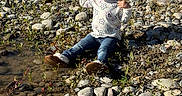 Avianna joined the competition — help win amazing prizes! toddler, child, sitting, rocks, riverbank, water, jeans, boots, long_sleeve_shirt, pigtails, curly_hair, outdoor, nature, sunlight, vegetation, distressed, expression, small_plant, ground, daylight