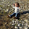 toddler, child, sitting, rocks, riverbank, water, jeans, boots, long_sleeve_shirt, pigtails, curly_hair, outdoor, nature, sunlight, vegetation, distressed, expression, small_plant, ground, daylight