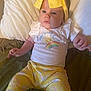 baby, child, headband, yellow_bow, infant, pillow, bed, clothing, cute, person, smiling_sun, rainbow, clouds, yellow_pants, patterned_clothing, indoors, portrait, lying_down, soft_light, happy