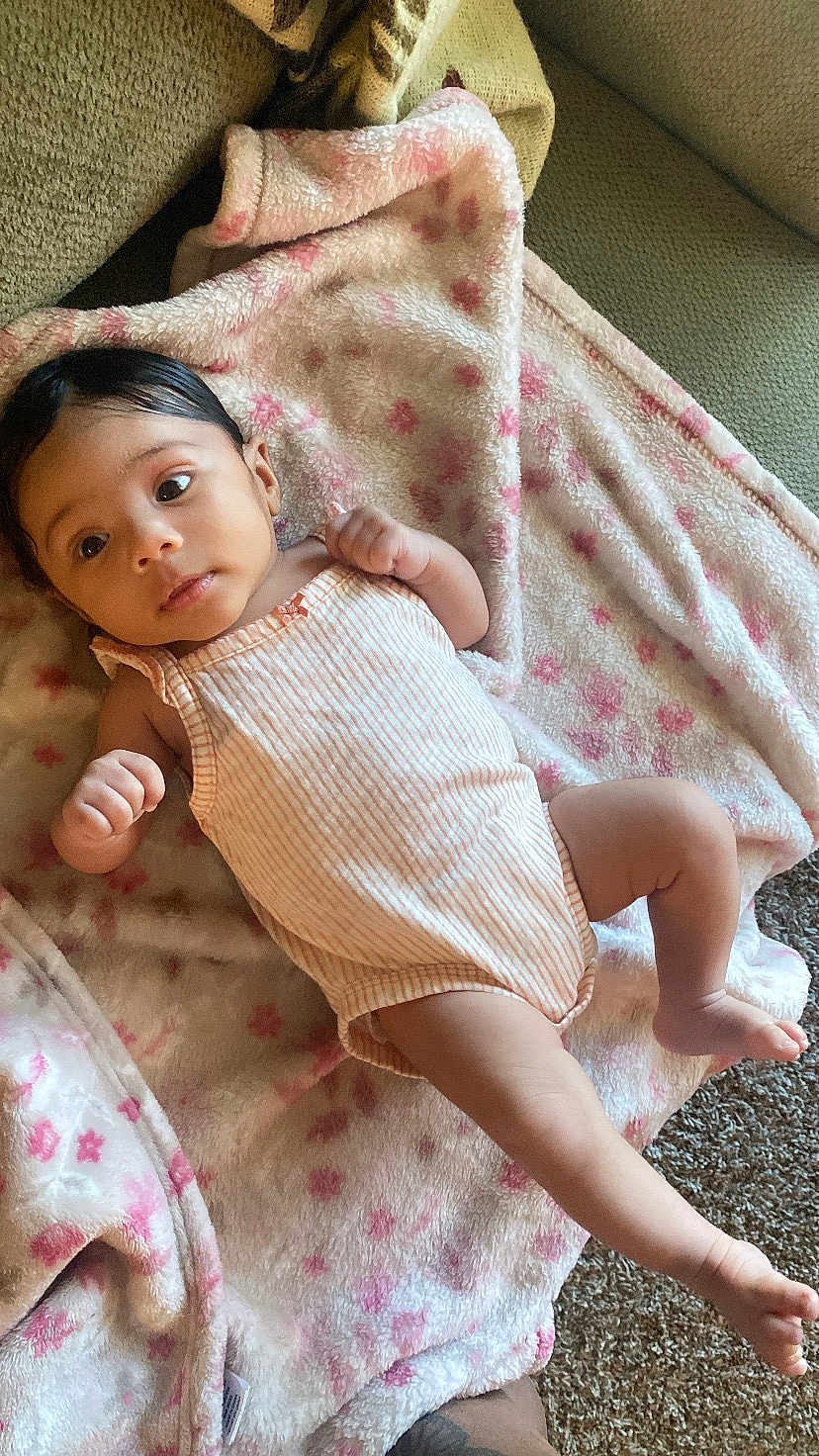 Amora joined the competition — help win amazing prizes! baby, infant, blanket, pink, striped_onesie, face, skin, hand, foot, lying_down, carpet, couch, soft_texture, child, newborn, indoors, relaxed, cute, small, person