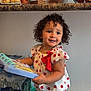 book, bottled_water, child, countertop, curly_hair, denim, eyes, fruit_bowl, hands, happy, indoor, kitchen, packaging, polka_dot, portrait, red_bow, refrigerator, smile, standing, toddler