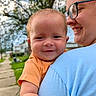 affection, baby, background_blur, blue_shirt, child, closeup, daylight, face, glasses, grass, happy, holding, nature, orange_shirt, outdoor, person, portrait, sidewalk, smile, tree