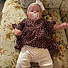 baby, pacifier, headband, polka_dot, maroon_top, white_pants, couch, floral_pattern, vintage, infant, lying_down, cute, clothing, indoor, child, furniture, portrait, one_arm_raised, soft_lighting, relaxed